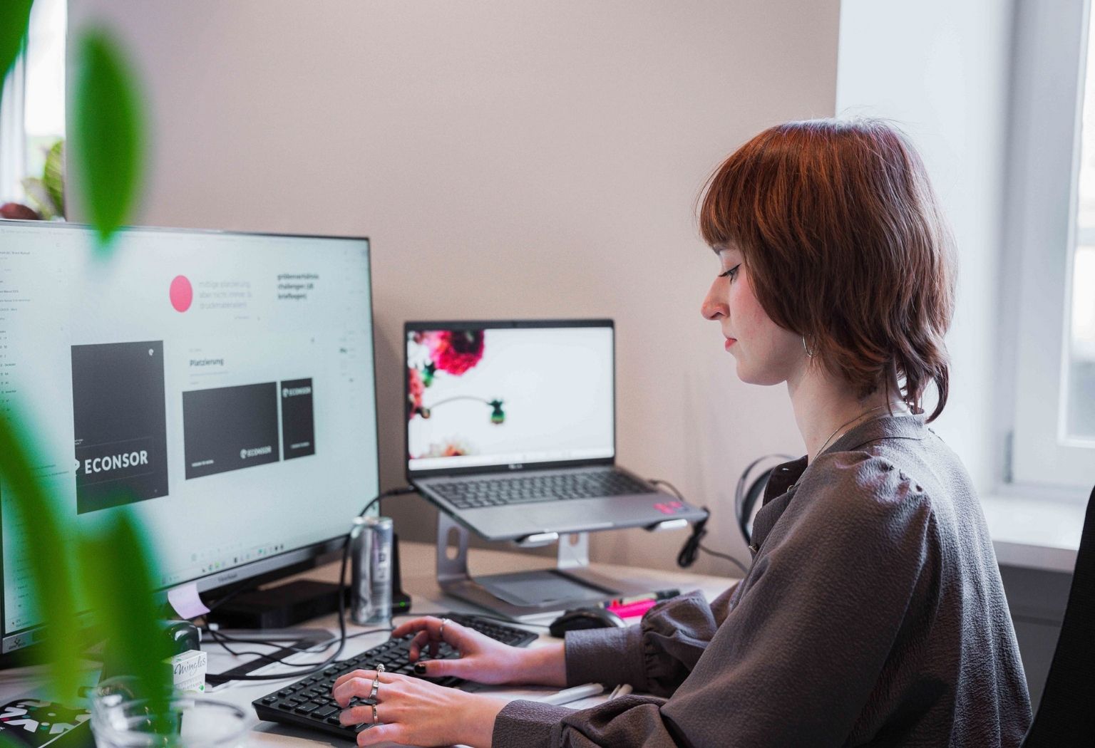 Eine Frau sitzt an einem Schreibtisch mit Computer und Monitor, vertieft in ihre Aufgaben in einem kreativen Arbeitsumfeld.